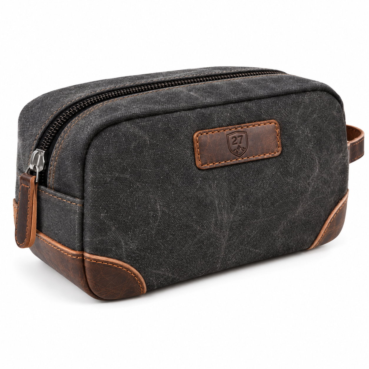 A rectangular zippered toiletry bag in dark gray waxed canvas with brown leather trim and corner protectors, featuring a stitched brand patch on the front.