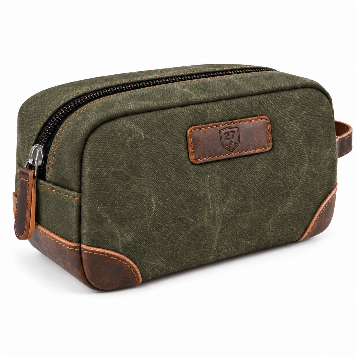 An olive green canvas toiletry bag with brown leather trim and a black zipper, featuring a brown leather patch with an embossed shield logo on the front.