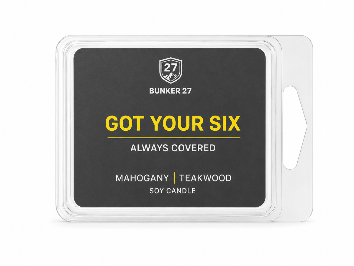 A clear plastic clamshell package containing a soy candle wax melt or melt wax product. The front label is dark charcoal gray with yellow and white text reading 'BUNKER 27', 'GOT YOUR SIX', 'ALWAYS COVERED', and 'MAHOGANY | TEAKWOOD SOY CANDLE'.